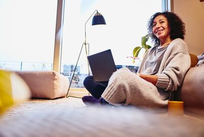 Smiling woman leaning against a couch, sitting on the floor, with laptop on her lap.