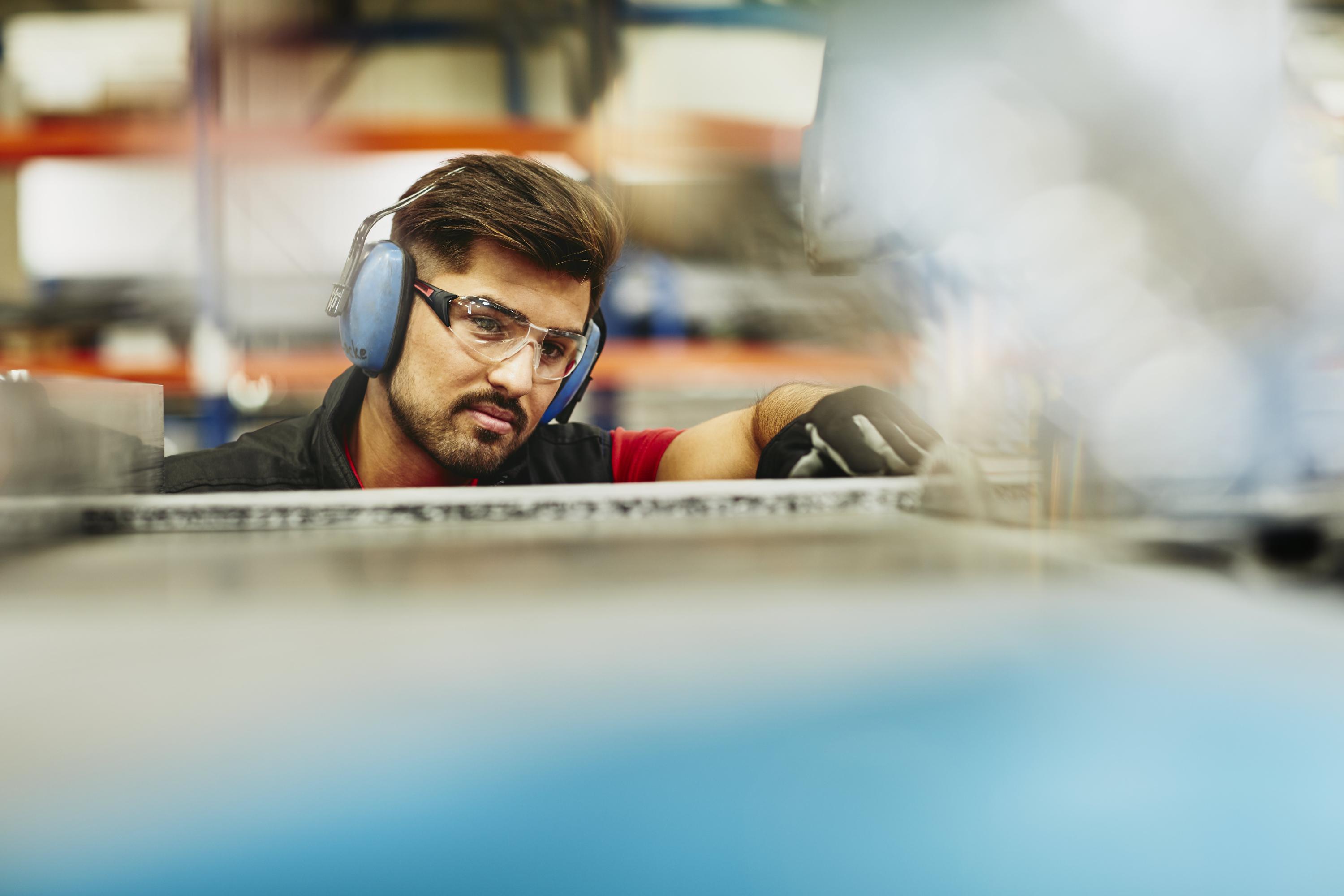 Male blue-collar worker in tech environment. Wearing protective ear covers, safety gloves. and safety goggles. Caucasian man. Groomed beard and mustache. Factory. Primary color blue. Secondary color cream.
